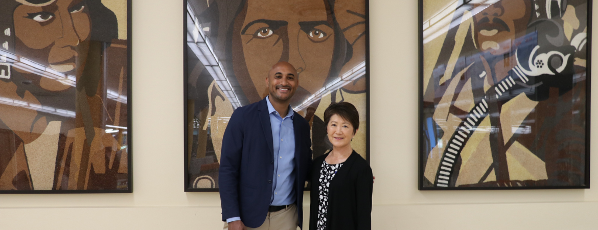 Congressman Shomari C. Figures and University Librarian Mihoko Hosoi standing together inside the UCR Tomás Rivera Library in front of three large paintings of Tuskegee Airmen, originally featured on a 2010 Rose Parade float.