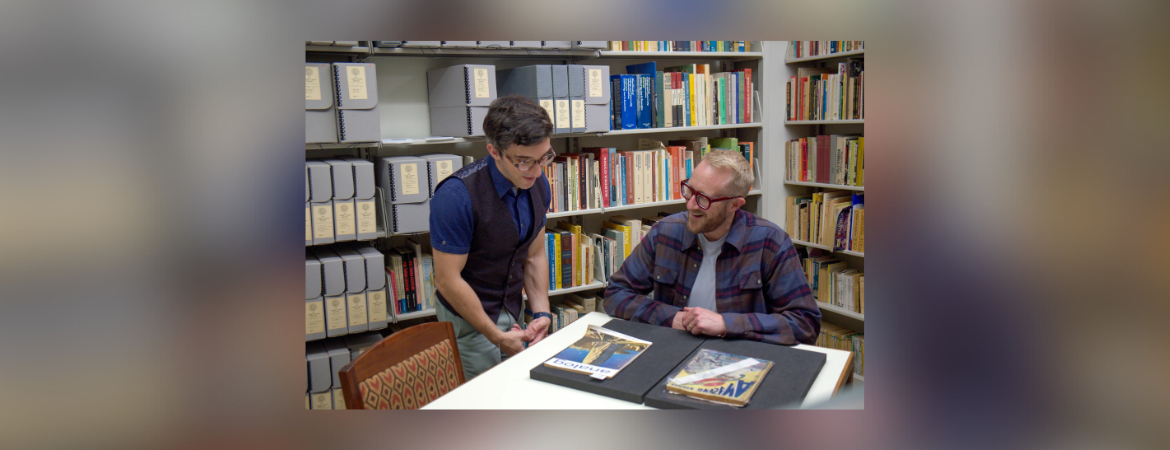 Two men examining documents at a table in a library with bookshelves in the background