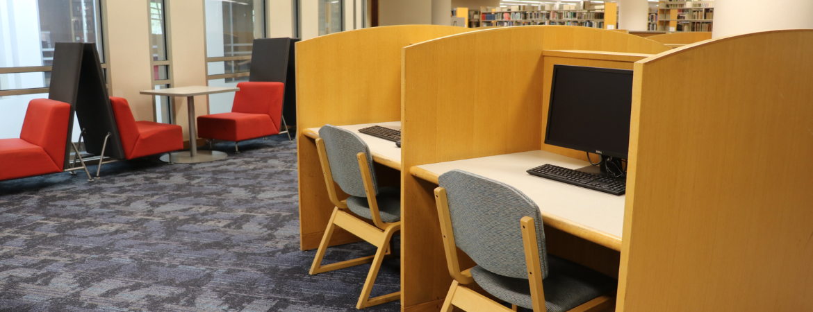A quiet, modern study area inside the Orbach Library, featuring rows of light-wood computer carrels with keyboards and monitors. To the left, there is a casual seating area with bright red chairs and tall privacy screens set against large windows, all atop a blue patterned carpet.