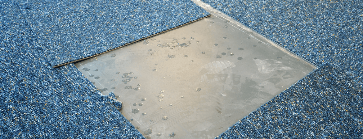 Blue speckled carpet tiles on a floor with one section removed, exposing the smooth gray subfloor and adhesive spots underneath.