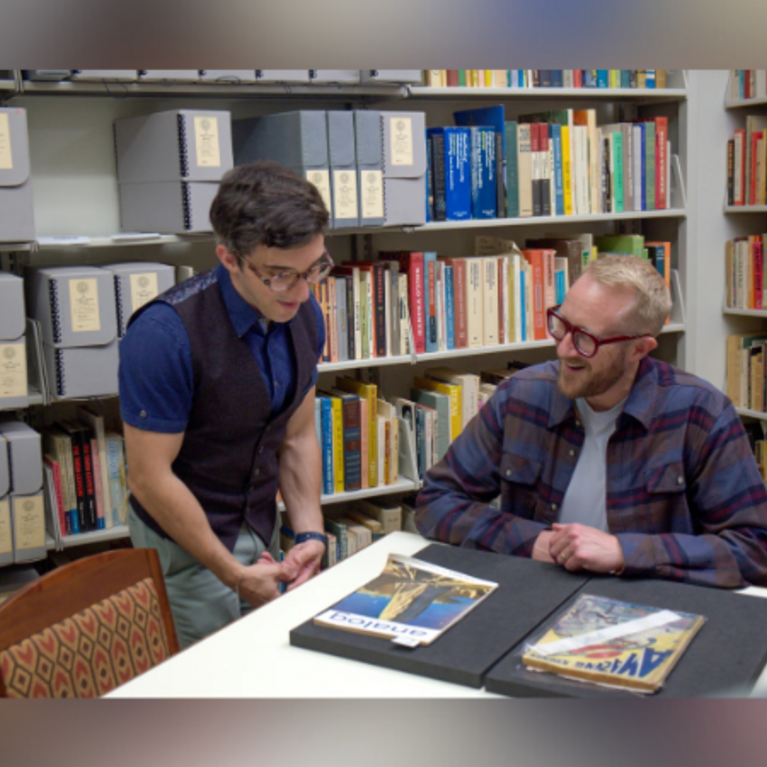 Two men examining documents at a table in a library with bookshelves in the background