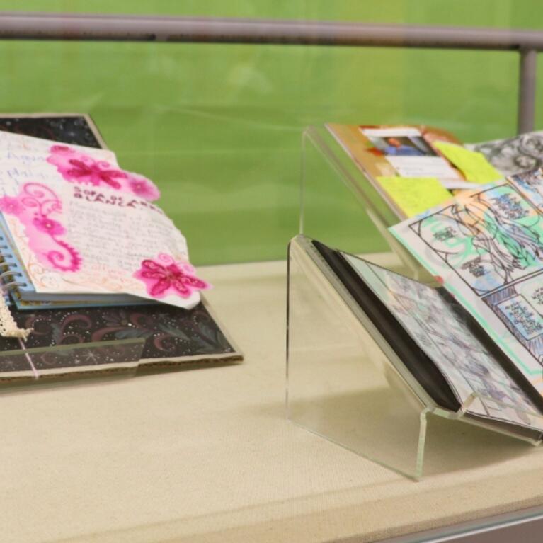A close-up view of a display case at the Rivera Library featuring open books with artistic "tippins." The books are layered with colorful mixed-media drawings, translucent Yupo paper, and personal ephemera as part of the ART 103 Ground Control exhibition.