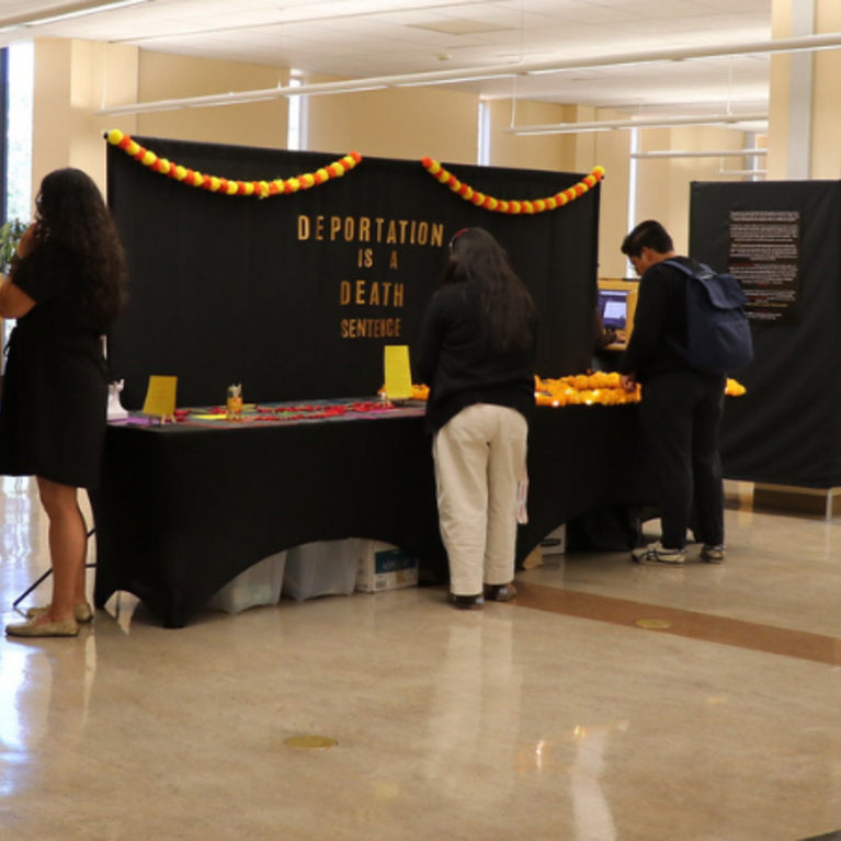 Visitors engage with the sensory stations of the "Amordidas Acompañadas" exhibit at UCR's Tomás Rivera Library. A black backdrop in the center reads "DEPORTATION IS A DEATH SENTENCE" in gold letters, decorated with orange marigold garlands.