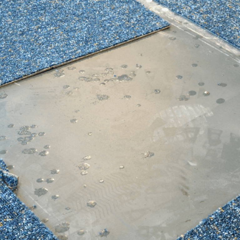 Blue speckled carpet tiles on a floor with one section removed, exposing the smooth gray subfloor and adhesive spots underneath.