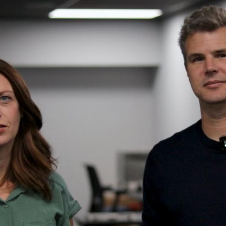 Rachael Faust in a green short-sleeved shirt and Mark Buchholz in a dark navy sweater stand side by side in a digitization lab, both wearing wireless lapel microphones.
