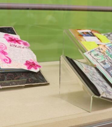 A close-up view of a display case at the Rivera Library featuring open books with artistic "tippins." The books are layered with colorful mixed-media drawings, translucent Yupo paper, and personal ephemera as part of the ART 103 Ground Control exhibition.