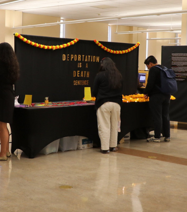Visitors engage with the sensory stations of the "Amordidas Acompañadas" exhibit at UCR's Tomás Rivera Library. A black backdrop in the center reads "DEPORTATION IS A DEATH SENTENCE" in gold letters, decorated with orange marigold garlands.