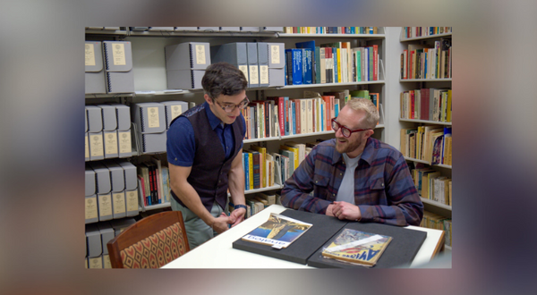 Two men examining documents at a table in a library with bookshelves in the background