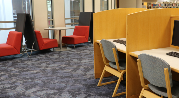 A quiet, modern study area inside the Orbach Library, featuring rows of light-wood computer carrels with keyboards and monitors. To the left, there is a casual seating area with bright red chairs and tall privacy screens set against large windows, all atop a blue patterned carpet.