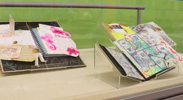A close-up view of a display case at the Rivera Library featuring open books with artistic "tippins." The books are layered with colorful mixed-media drawings, translucent Yupo paper, and personal ephemera as part of the ART 103 Ground Control exhibition.