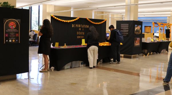 Visitors engage with the sensory stations of the "Amordidas Acompañadas" exhibit at UCR's Tomás Rivera Library. A black backdrop in the center reads "DEPORTATION IS A DEATH SENTENCE" in gold letters, decorated with orange marigold garlands.