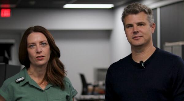 Rachael Faust in a green short-sleeved shirt and Mark Buchholz in a dark navy sweater stand side by side in a digitization lab, both wearing wireless lapel microphones.