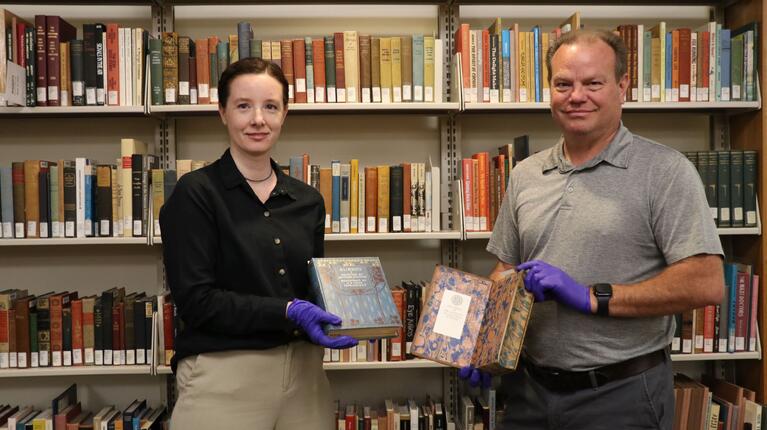UCR Library’s Christina Bean and EH&S’s Mark Atwood standing in the library stacks. Both are wearing purple nitrile gloves and holding open Victorian-era books with decorative bindings to demonstrate safe handling procedures.
