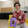 UCR Library's Dr. Phoenix Alexander smiles and holds up a copy of The Dream Hotel by Laila Lalami while seated in a yellow armchair.