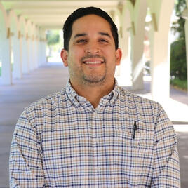 Portrait of a smiling man with short dark hair and light facial hair, wearing a white, blue, and tan plaid button-down shirt with a pen in his front pocket, outdoors in an arched walkway.